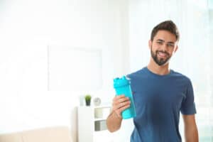 A man with a beard and a blue shirt holds a blue shaker bottle. He is standing indoors in a bright room, embodying stress resilience and ANS balance.