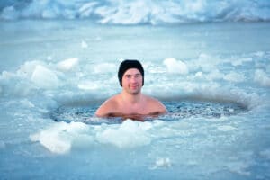A man wearing a black beanie is immersed in icy water, with ice and snow surrounding the hole he is in, demonstrating remarkable stress resilience.